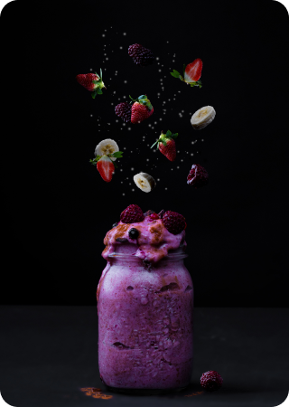 fotografia de um vidro cheio de shake roxo com frutas espalhadas no ar logo acima dele